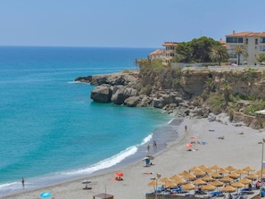 A coastal beach with clear blue waters, rocky cliffs, and a row of beach umbrellas. - Spectacular front sea view apartment (Torrox Costa)