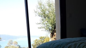 A bedroom with a bed, a large window, and a view of a body of water and greenery. - Exceptional house in quiet magnificent view (Olmeto)
