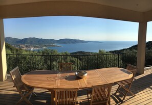 Une terrasse en bois avec une table ronde et des chaises, offrant une vue sur un paysage côtier avec des montagnes et la mer.