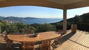 A wooden deck with a round table and chairs, overlooking a coastal landscape with mountains and a body of water. - Exceptional house in quiet magnificent view (Olmeto)