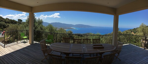 Un espace extérieur abrité, avec une table et des chaises en bois, une chaise verte, et une vue sur la mer et les montagnes.