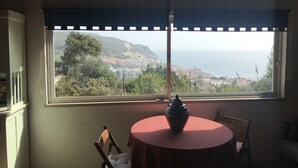 A dining area with a table and chairs, a view of a coastal town, and a window with a view of the sea and hills.