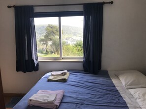 A room with a bed, a window with curtains, and a view of greenery and buildings outside.