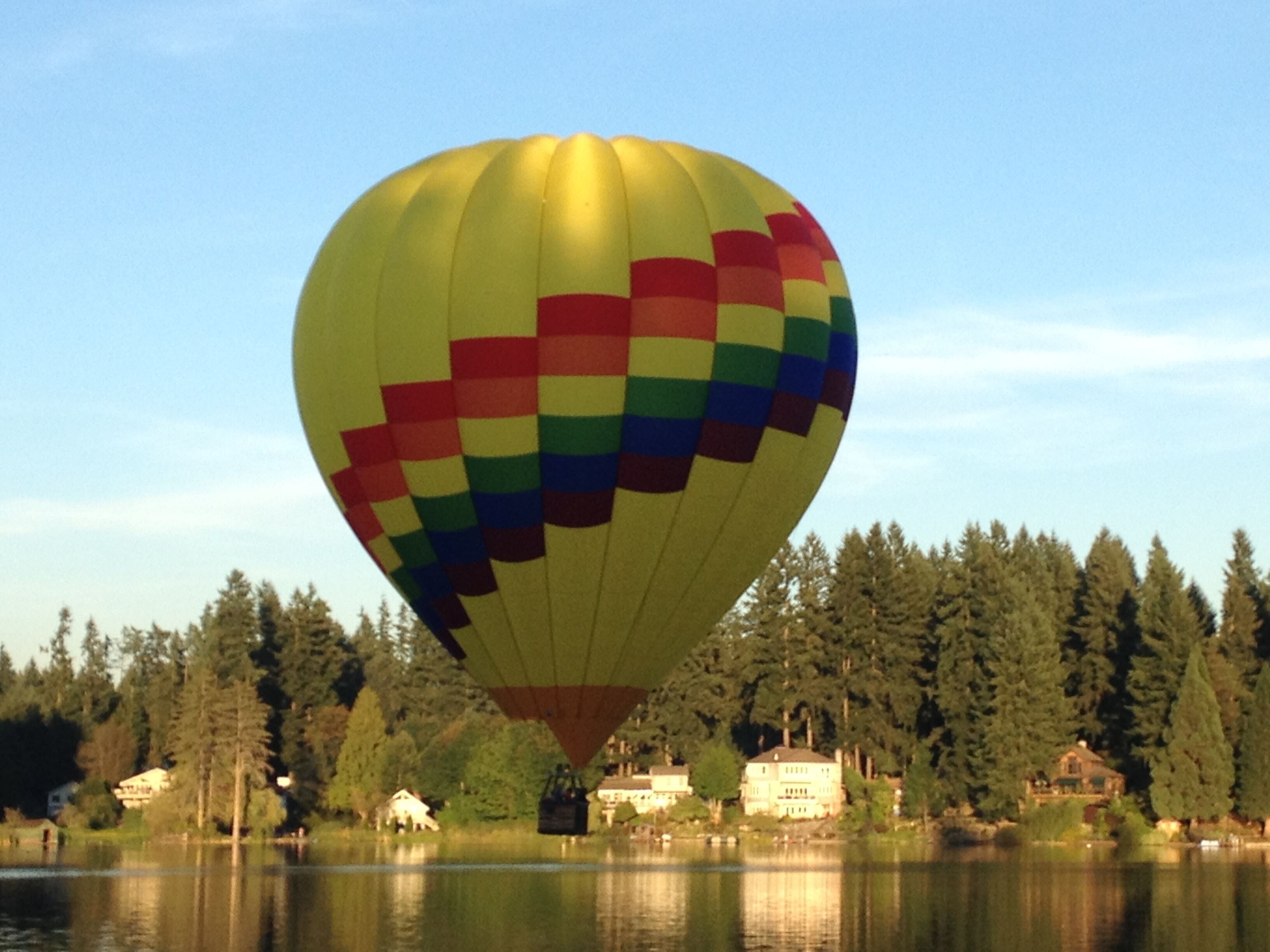 A hot air balloon with a rainbow pattern flying over a lake with houses and trees in the background.