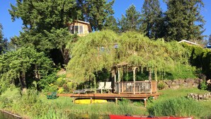 A wooden deck with chairs, a small gazebo, and a red canoe on a calm body of water, surrounded by lush greenery and trees. - Cottage Lake Bed and Breakfast (Woodinville)