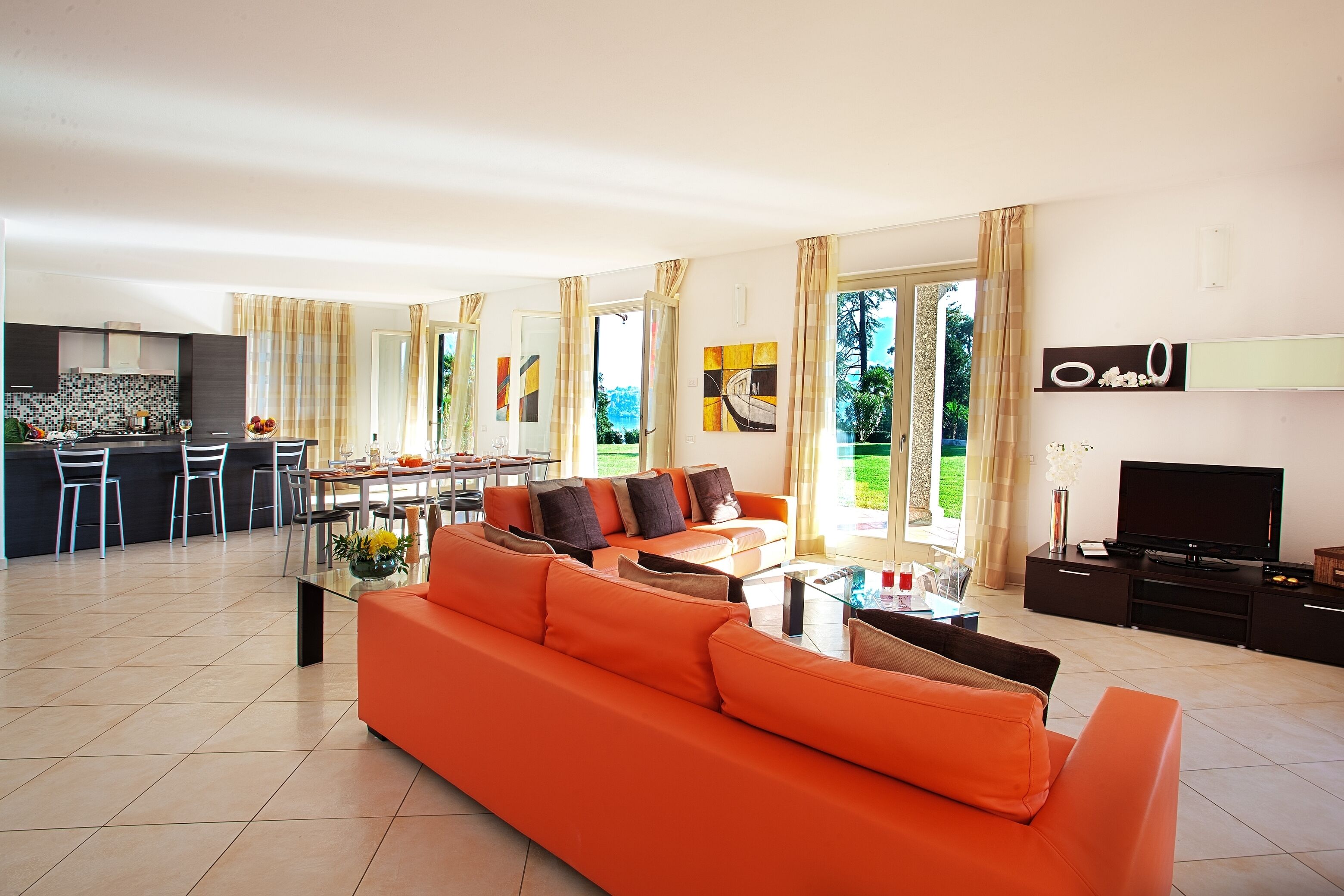 Una sala moderna con un sofá naranja grande, un comedor con mesa y sillas, una cocina con armarios oscuros y un televisor sobre un mueble oscuro.
