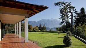 A covered outdoor area with a tiled floor, surrounded by a well-maintained lawn and trees, with mountains in the background.