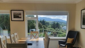 A dining area with a wooden table and chairs, a black chair with a yellow cushion, and a view of greenery and mountains through large windows.
