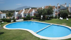 A swimming pool with a curved edge, surrounded by a well-maintained lawn and white buildings with red-tiled roofs. - TOWN HOUSE IN SECURE  URBANIZATION.   AIR CON.   INTERNET.   MOSQUITO SCREENS. (ESTEPONA)