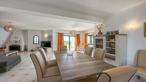 Un vaste salon doté d’une cheminée, un coin repas avec une longue table et des chaises, et une cuisine équipée d’un meuble et d’étagères.