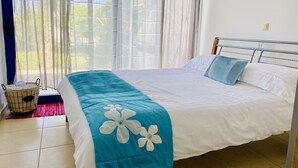 Ein ordentlich bezogenes Bett mit Blumenmuster, einem Metall-Kopfschild und einem Fenster mit durchsichtigen Vorhängen.