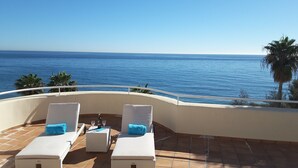 A terrace with two white cushioned lounge chairs, a small table with a bottle and glasses, and a view of the ocean.