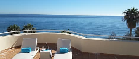 A terrace with two white cushioned lounge chairs, a small table with a bottle and glasses, and a view of the ocean.