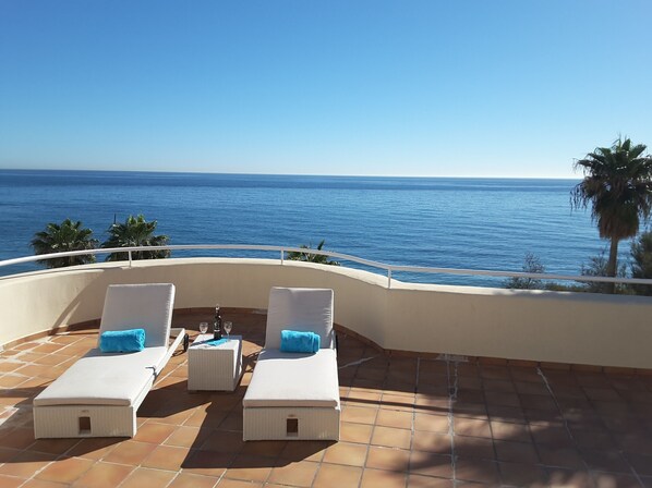 A terrace with two white cushioned lounge chairs, a small table with a bottle and glasses, and a view of the ocean.