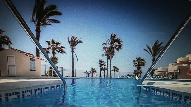 A swimming pool with clear blue water, surrounded by palm trees and a building with a beige facade.