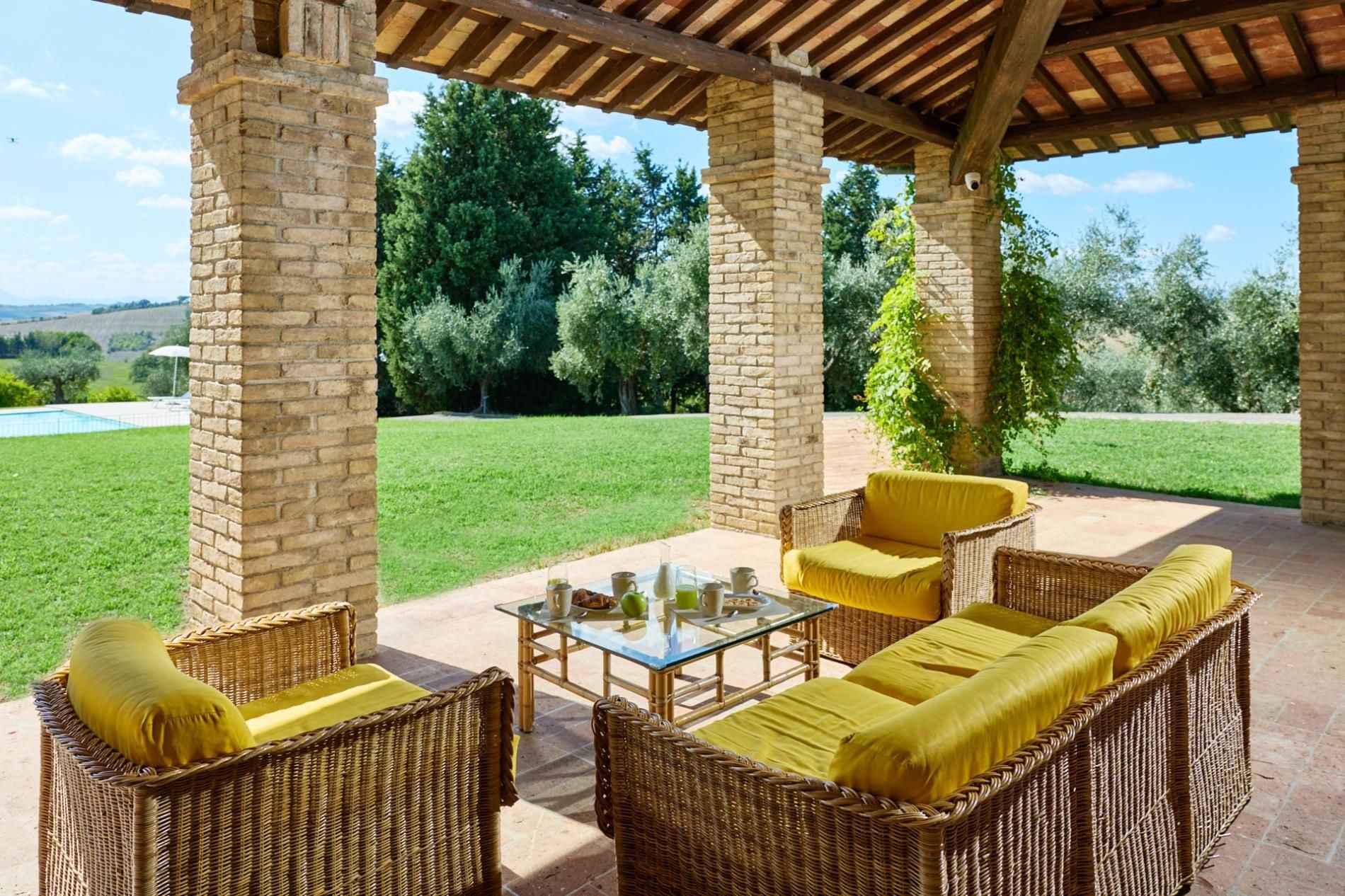 Une terrasse extérieure aménagée avec des meubles en osier et une table en verre, entourée de piliers en briques et offrant une vue sur un paysage verdoyant.