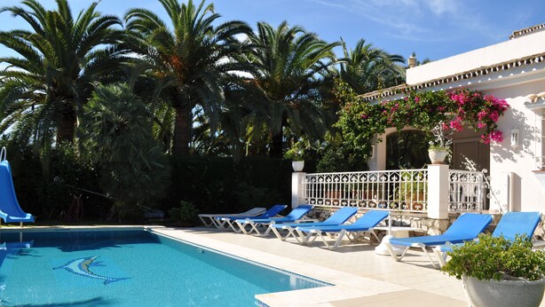 A swimming pool with blue lounge chairs and a white building with a decorative archway.