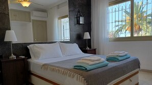 A bedroom with a bed, two bedside tables, a ceiling fan, a window with a view of palm trees, and a mirror on the wall.