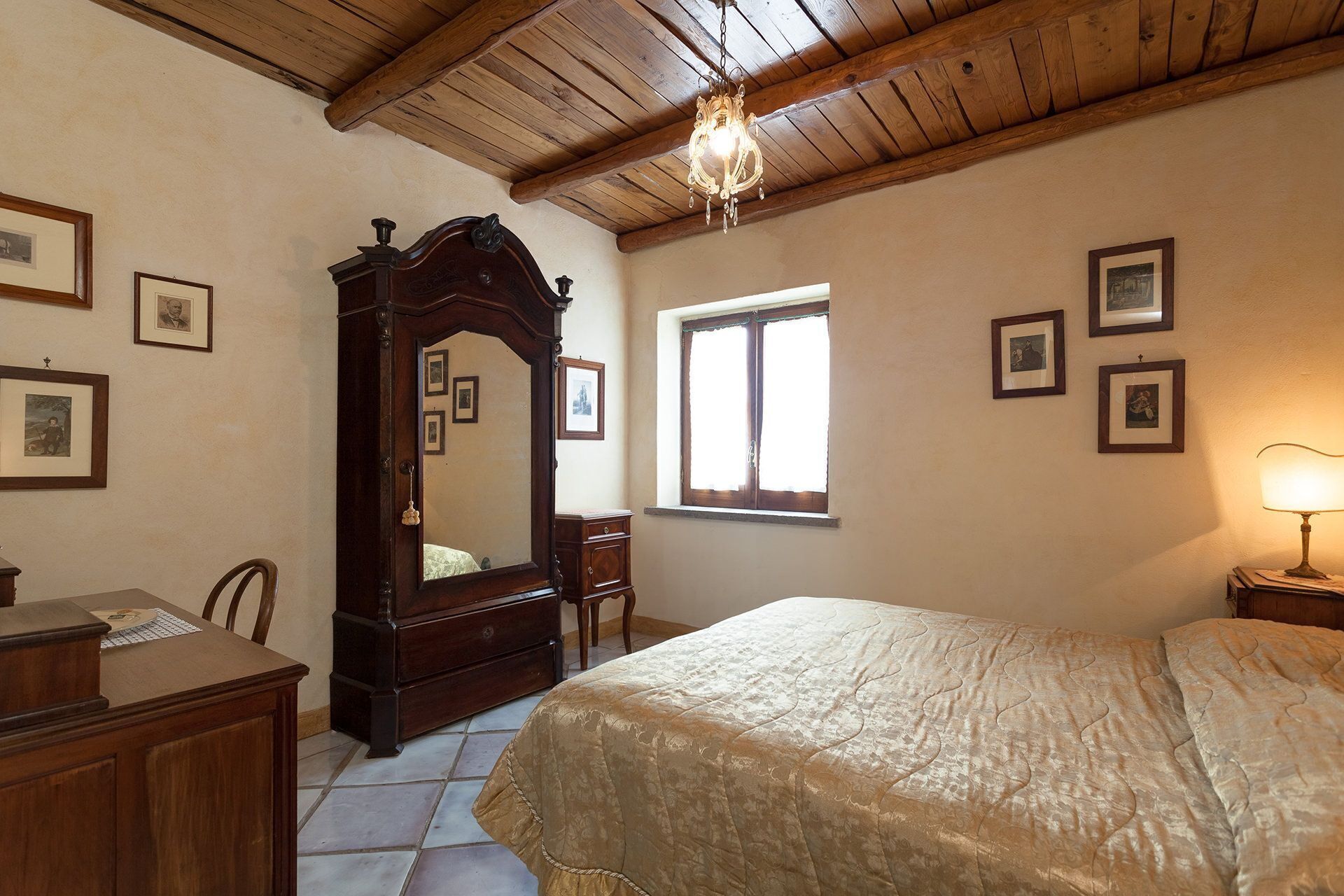 A bedroom with a wooden ceiling, a large mirror, a bed with a beige bedspread, a desk with a chair, and framed pictures on the walls.