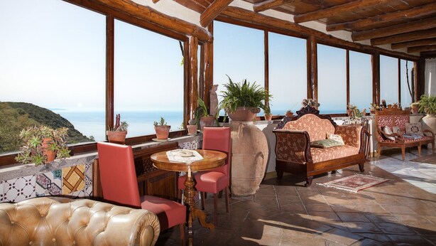 A spacious terrace with a wooden ceiling, large windows offering a view of the sea, and various seating arrangements including a sofa, chairs, and a table.