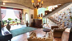 A spacious living room with a wooden floor, a chandelier, a staircase with a mosaic wall, and a view of the garden through a large window.