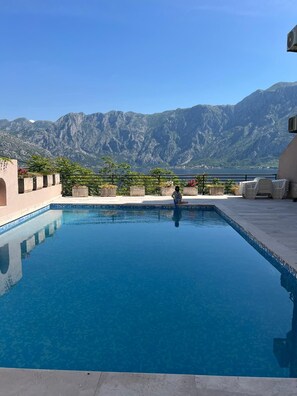 A rectangular pool with clear blue water, surrounded by a low stone wall and potted plants. In the background, there are mountains and a clear sky.