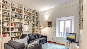 A living room with a grey sofa, a bookshelf, a TV, and a window. - 3 Bdr. Apt. Near Piazza Navona | 4 AC, WiFi, Heat (Roma)