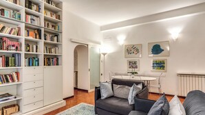 A living room with a bookshelf, a gray sofa, a dining table, and artwork on the wall. - 3 Bdr. Apt. Near Piazza Navona | 4 AC, WiFi, Heat (Roma)