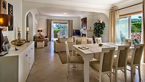 Un grand espace de vie comprenant une table à manger, des canapés et un téléviseur. Un balcon offre une vue sur la verdure.
