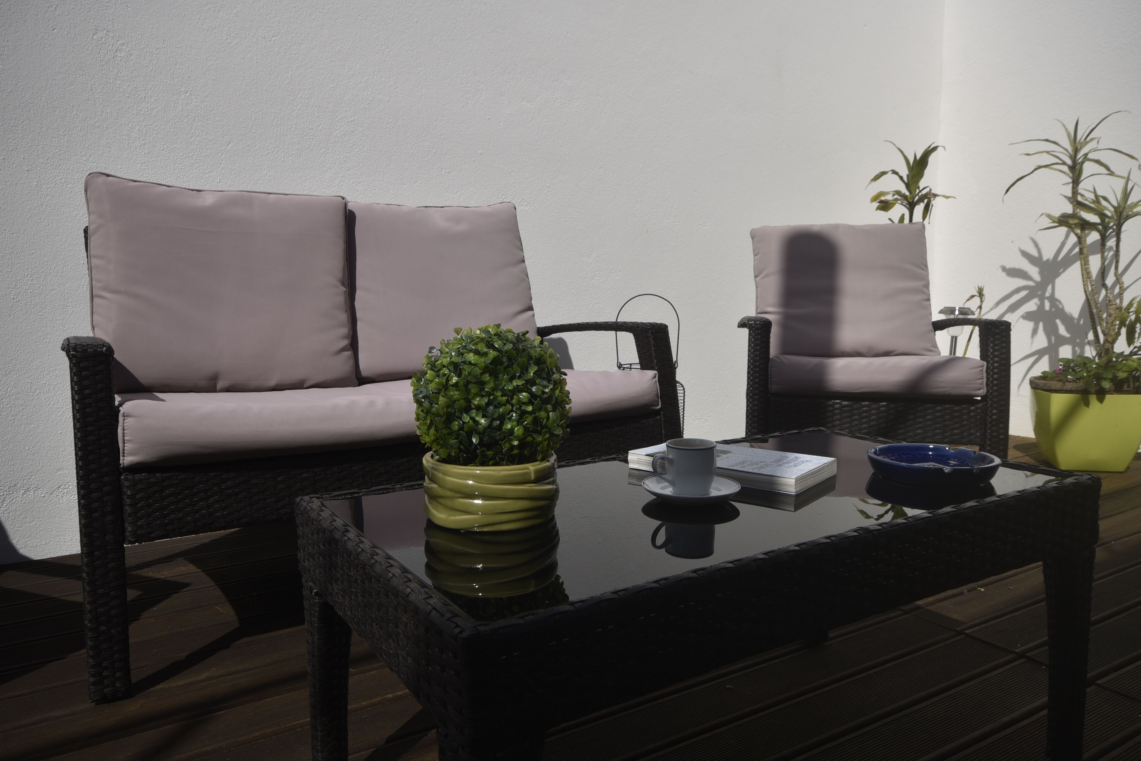 Un área de estar con dos sillas de mimbre y una mesa, una planta en maceta y una mesita con una taza y un cuenco.