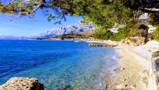 A coastal beach with clear blue waters, rocky shoreline, and a backdrop of mountains. - Apartment with stunning sea views (Podgora)