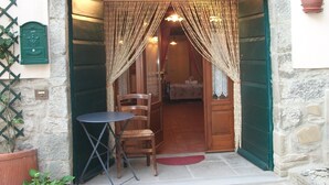 A stone building entrance with a wooden door, a small table with a chair, and a potted plant on the right. - Typical Tuscan style apartment (cortona)