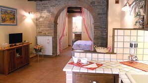 A rustic room with a stone archway leading to a bedroom, a wooden cabinet with a TV, and a tiled bathroom area. - Typical Tuscan style apartment (cortona)