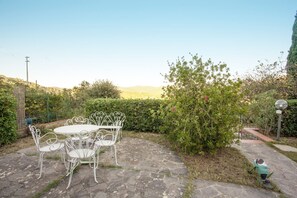 A stone patio with a round table and white metal chairs, surrounded by greenery and a view of hills.