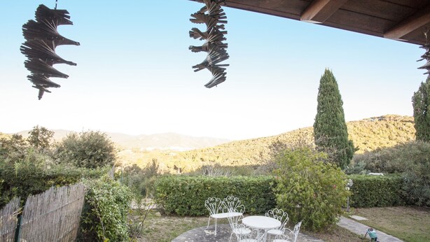 A patio with white metal chairs and a table, surrounded by greenery and a view of hills. - VILLETTA BEL GIARDINO (MARINA DI CAMPO)