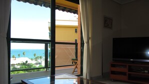 Een woonkamer met een groot raam dat uitzicht biedt op de oceaan en palmbomen. Er staat een flatscreen televisie op een houten voet.