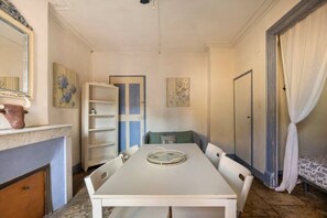 A dining area with a white table, chairs, and a decorative plate. There is a white cabinet with a vase on top, a bookshelf, and a bed with a patterned bedspread.