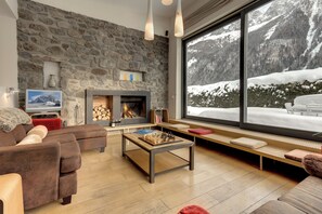 A cozy living room with a stone wall, a fireplace, a large window with a snowy mountain view, a sofa, and a coffee table with chess pieces.