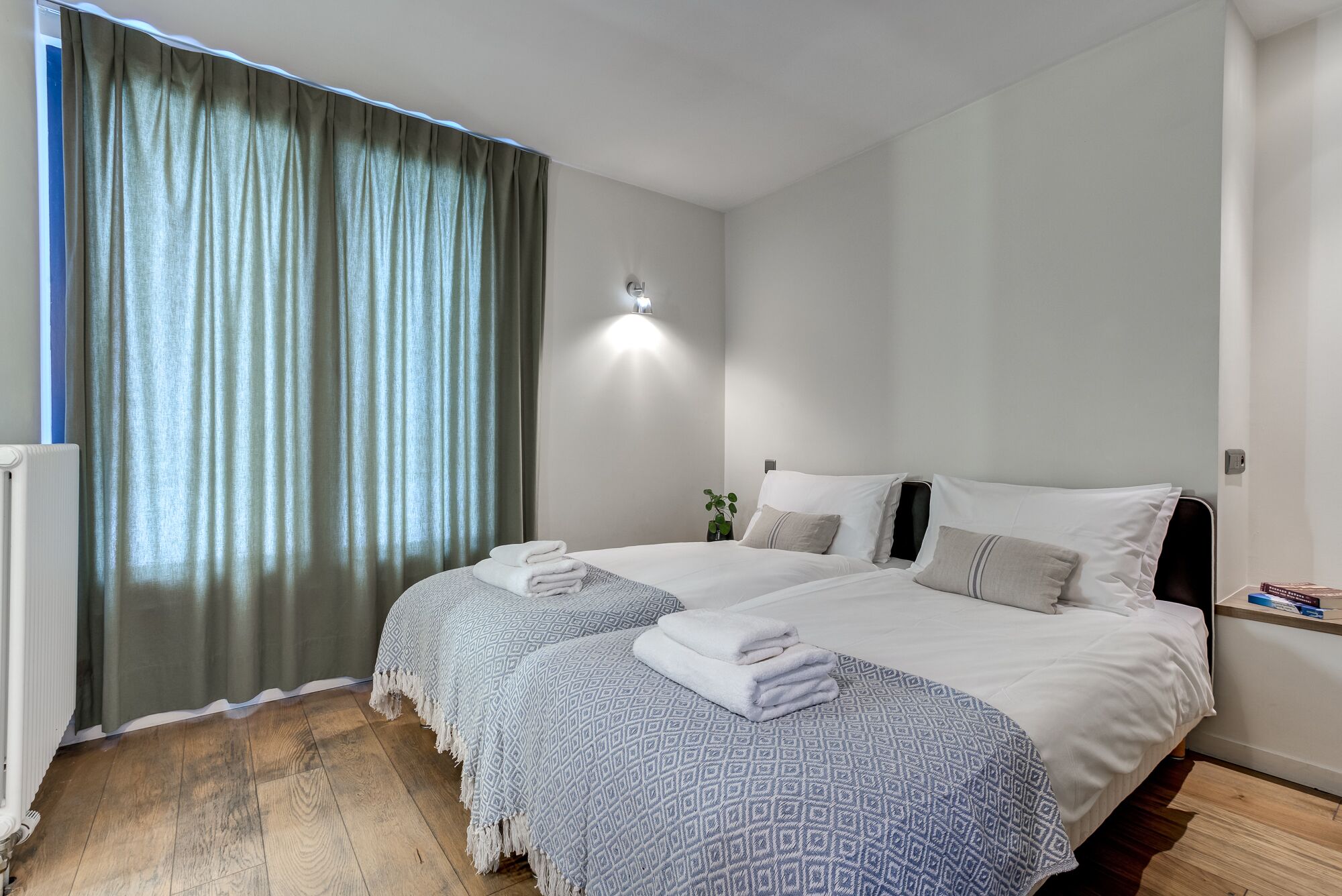 A hotel room with two beds, a large window with curtains, a wall-mounted light, and a radiator.
