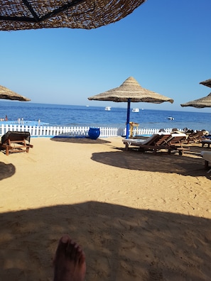 A beach with sun loungers and thatched umbrellas, a white fence, and boats in the water.