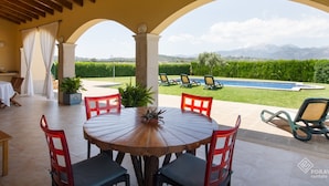 Un porche techado con una mesa redonda de madera y sillas rojas, con vistas a una piscina y montañas.