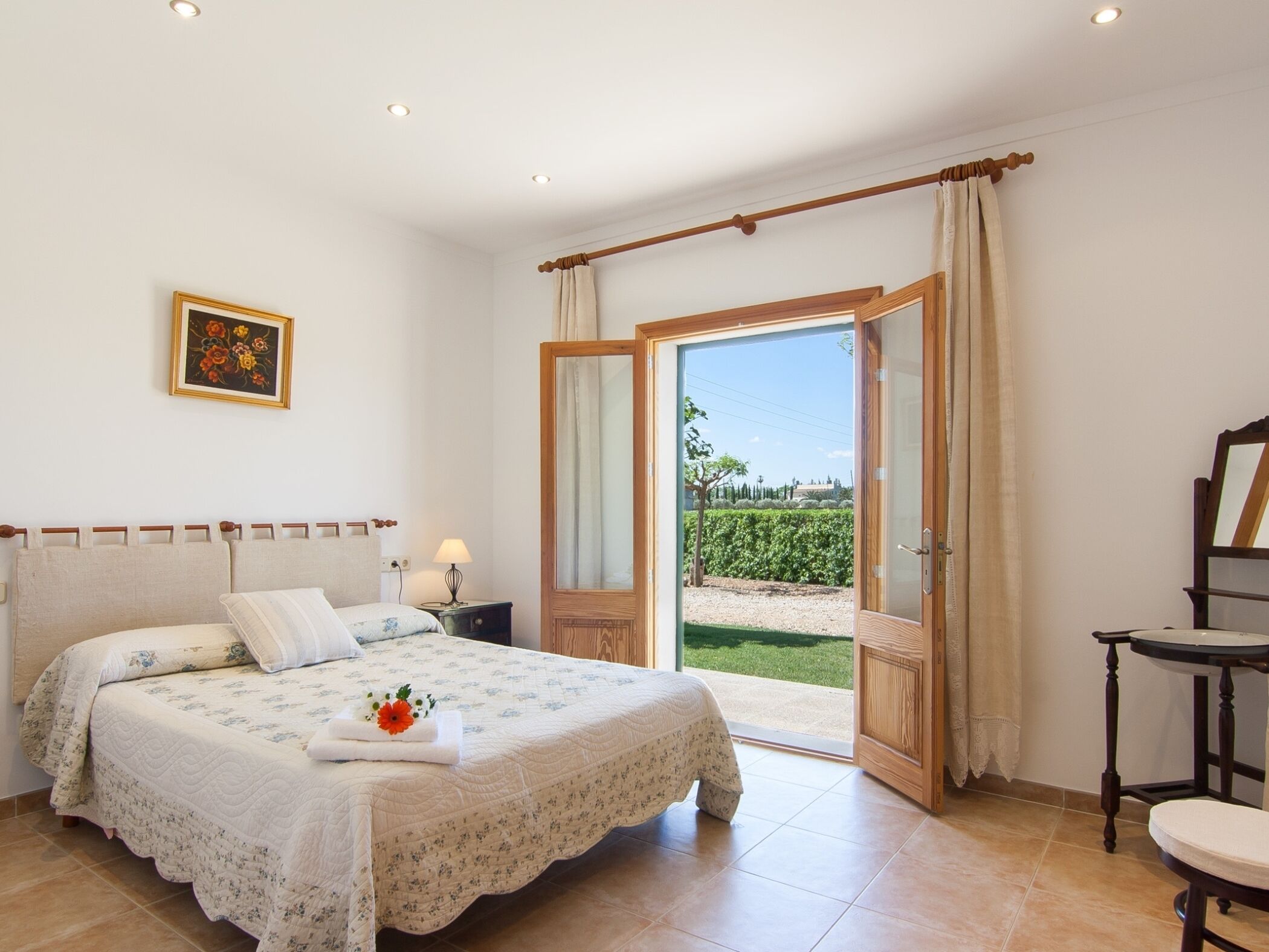 A bedroom with a bed, a nightstand, a chair, and a view of a garden through an open door.