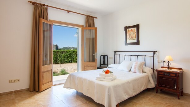 Un dormitorio con cama, mesita de noche, lámpara, un cuadro en la pared y vistas al exterior.
