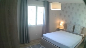 Um quarto com uma cama, uma mesa de cabeceira com abajur, uma janela com cortinas e uma parede estampada.