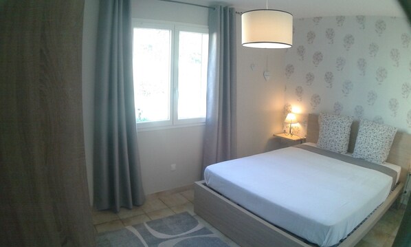 A bedroom with a bed, a nightstand with a lamp, a window with curtains, and a patterned wall.
