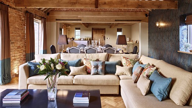 A cozy living room with a high ceiling, exposed wooden beams, a brick wall, and a large beige sofa with multiple cushions.