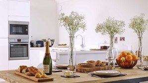 Una cocina moderna con una mesa de madera dispuesta para una comida, mostrando pasteles, una botella de champán y flores decorativas.