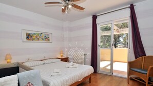 Quarto com cama, cadeira, ventoinha no teto, mesa de cabeceira, abajur, janela com cortinas e um quadro na parede.