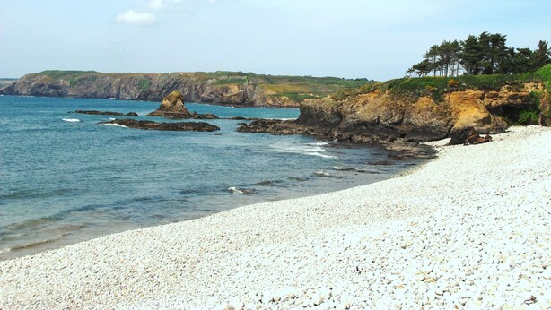 Una playa de guijarros con afloramientos rocosos y un acantilado cubierto de hierba.