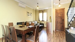 Uma área de refeições com mesa e cadeiras de madeira, um sofá e uma escada que dá acesso a um andar superior.